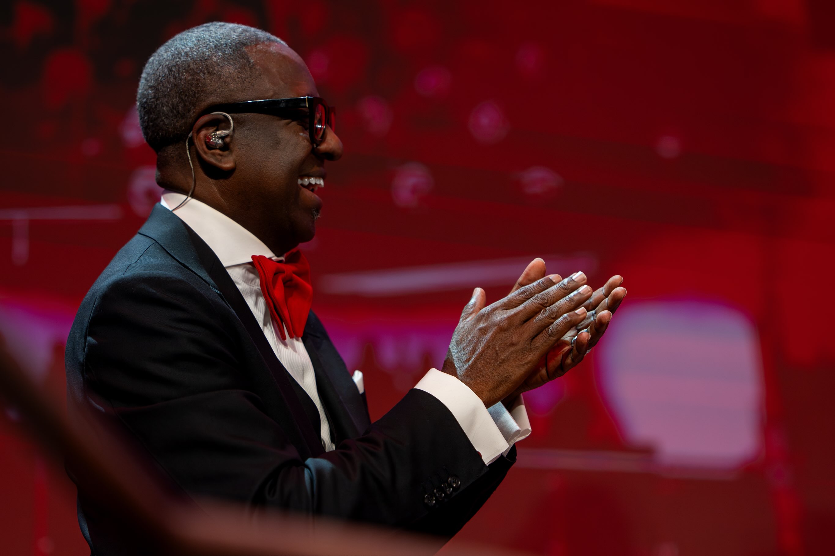 Minister Leon C. Lewis, silver-haired and smiling, clapping in a black tuxedo and red bow tie against a red-lit stage